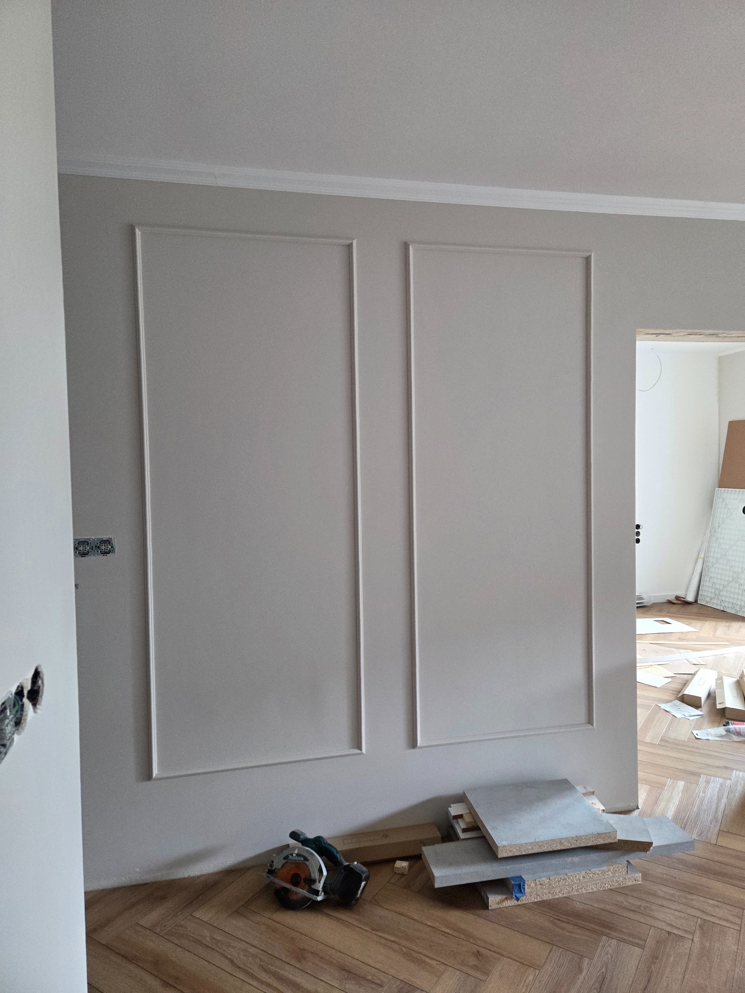 Ściana z dekoracyjnymi panelami w stylu klasycznym, świeżo po montażu. Na podłodze leżą narzędzia i fragmenty paneli. Widoczny parkiet w jodełkę i fragment instalacji elektrycznej.