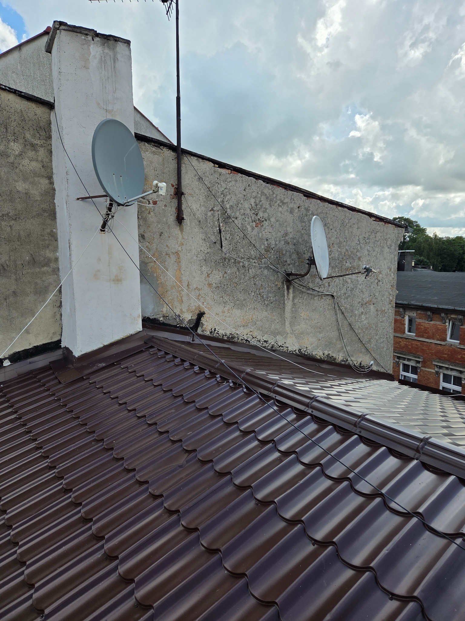 Nowy, brązowy dach z blachy trapezowej, widok z góry. Dwie anteny satelitarne na tle budynku z szarą elewacją i fragmentem nieba z chmurami.