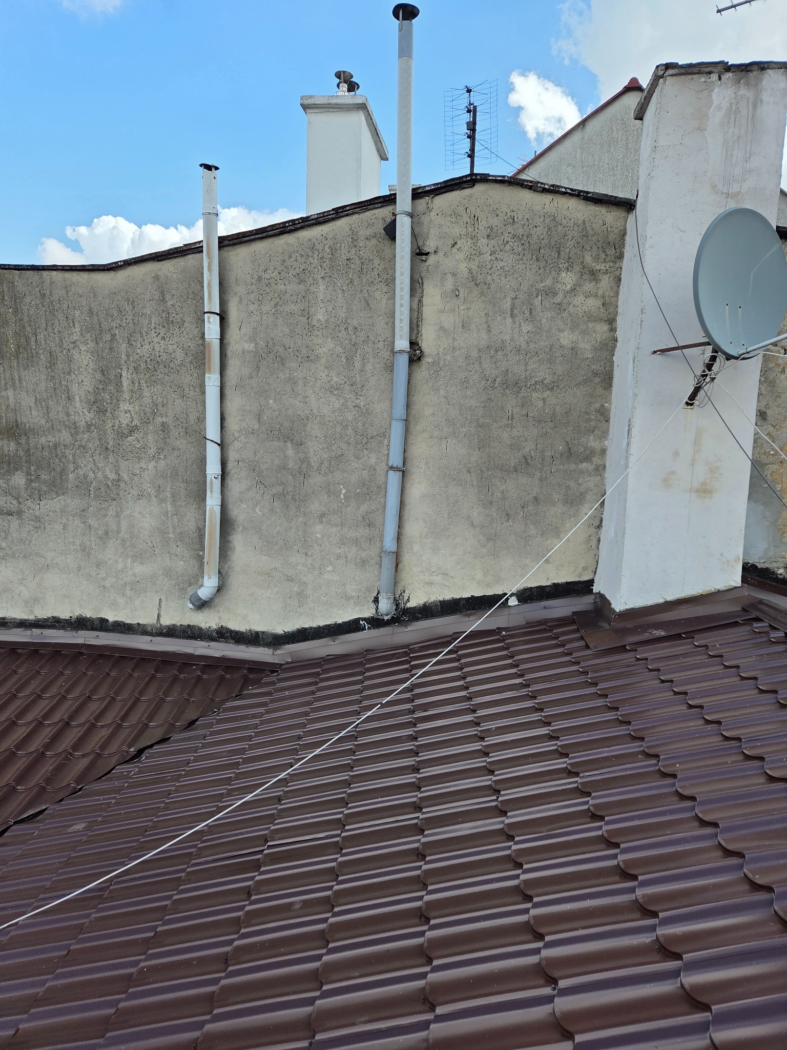 Brązowy dach z blachy trapezowej na tle szarej ściany budynku z kominami i anteną satelitarną. Widok z góry, w słoneczny dzień.