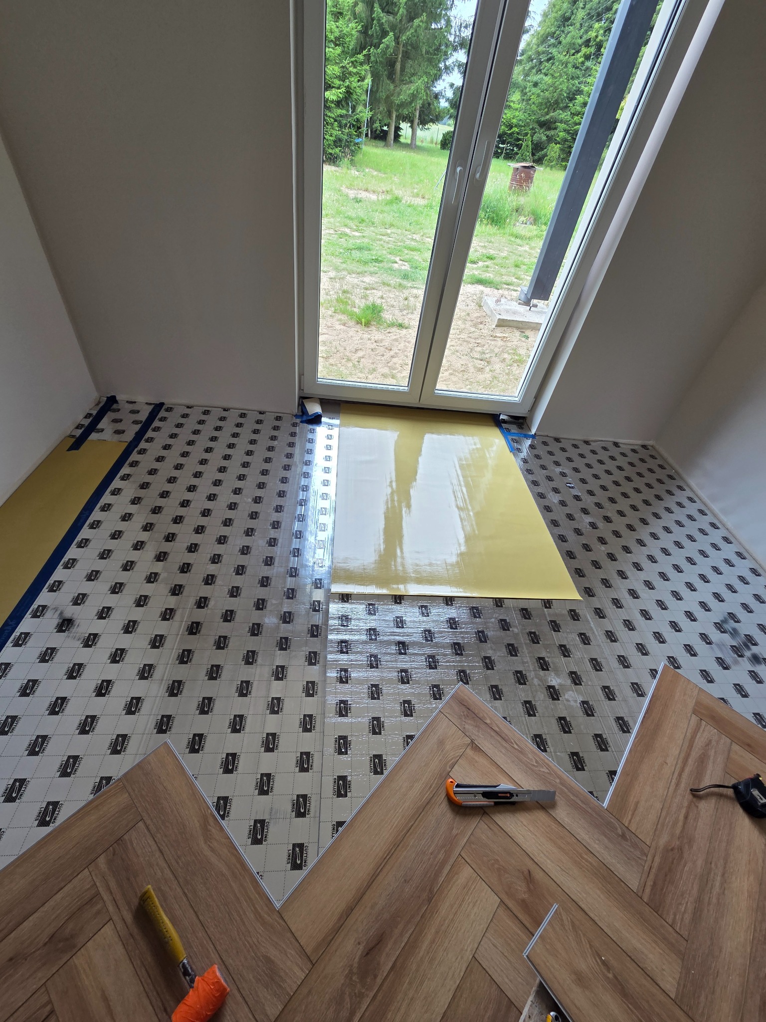 Układanie paneli podłogowych w jodełkę w pokoju na podkładzie izolacyjnym. Widoczne narzędzia: nóż, młotek. Okno dachowe w tle. Częściowo ułożona podłoga.