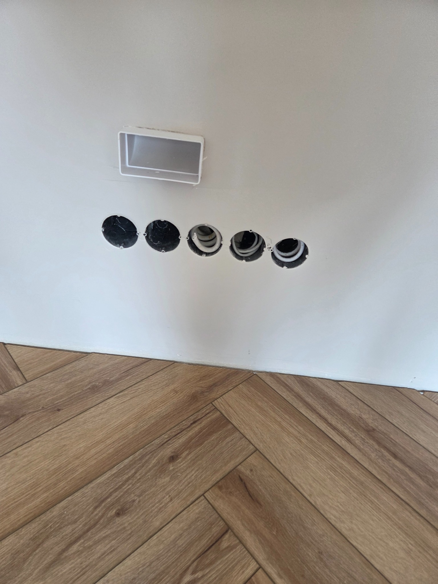 Biała ściana z pięcioma okrągłymi otworami na kable i puszką instalacyjną, widoczne przewody, parkiet ułożony w jodełkę na podłodze, ujęcie z dołu.