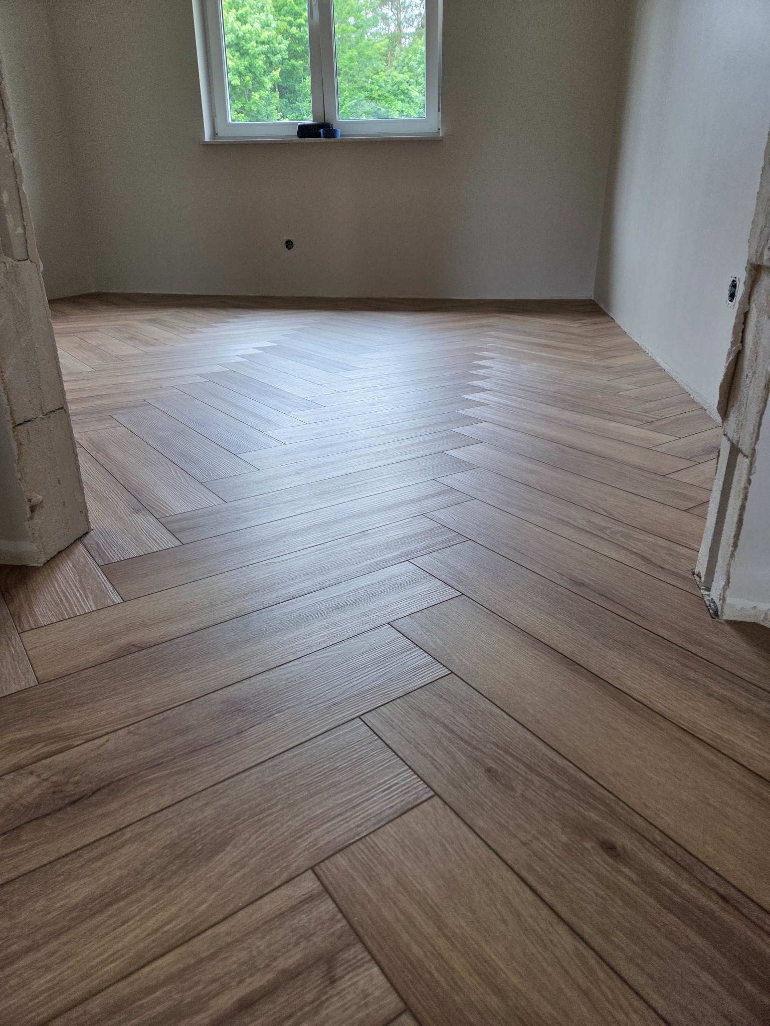 Ukończona podłoga z paneli ułożonych w jodełkę w Świdwinie. Widok na jasne wnętrze z oknem i fragmentem surowej ściany. Nowoczesny design.