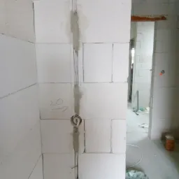 Białe bloczki ścienne z wyżłobionym pionowym rowkiem wypełnionym szarym cementem, w którym umieszczono przewody elektryczne; widoczny otwór na puszkę elektryczną z wystającymi kablami oraz napis...