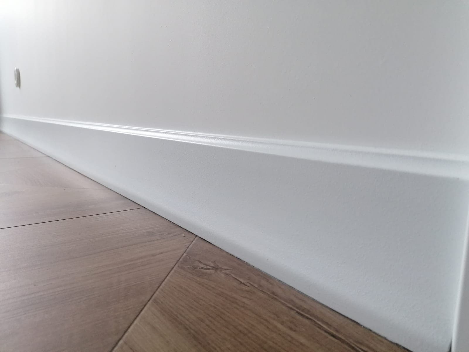 Biała listwa przypodłogowa zamontowana przy ścianie na drewnianej podłodze. Detal wykończenia wnętrza, czysty montaż, subtelne oświetlenie.