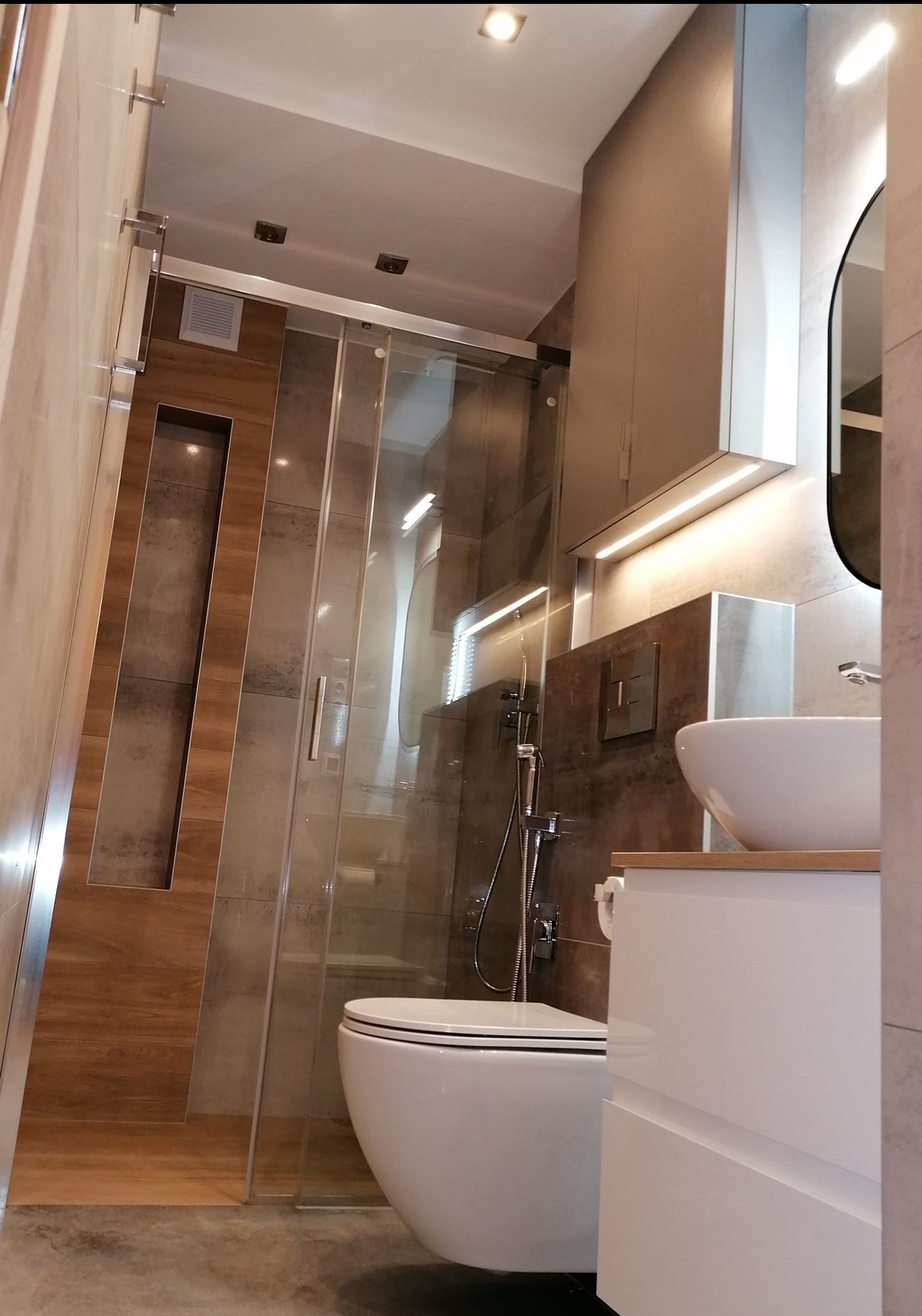 Nowoczesna łazienka z kabiną prysznicową typu walk-in, umywalką nablatową i drewnianymi akcentami. Widoczne oświetlenie LED i minimalistyczna armatura.