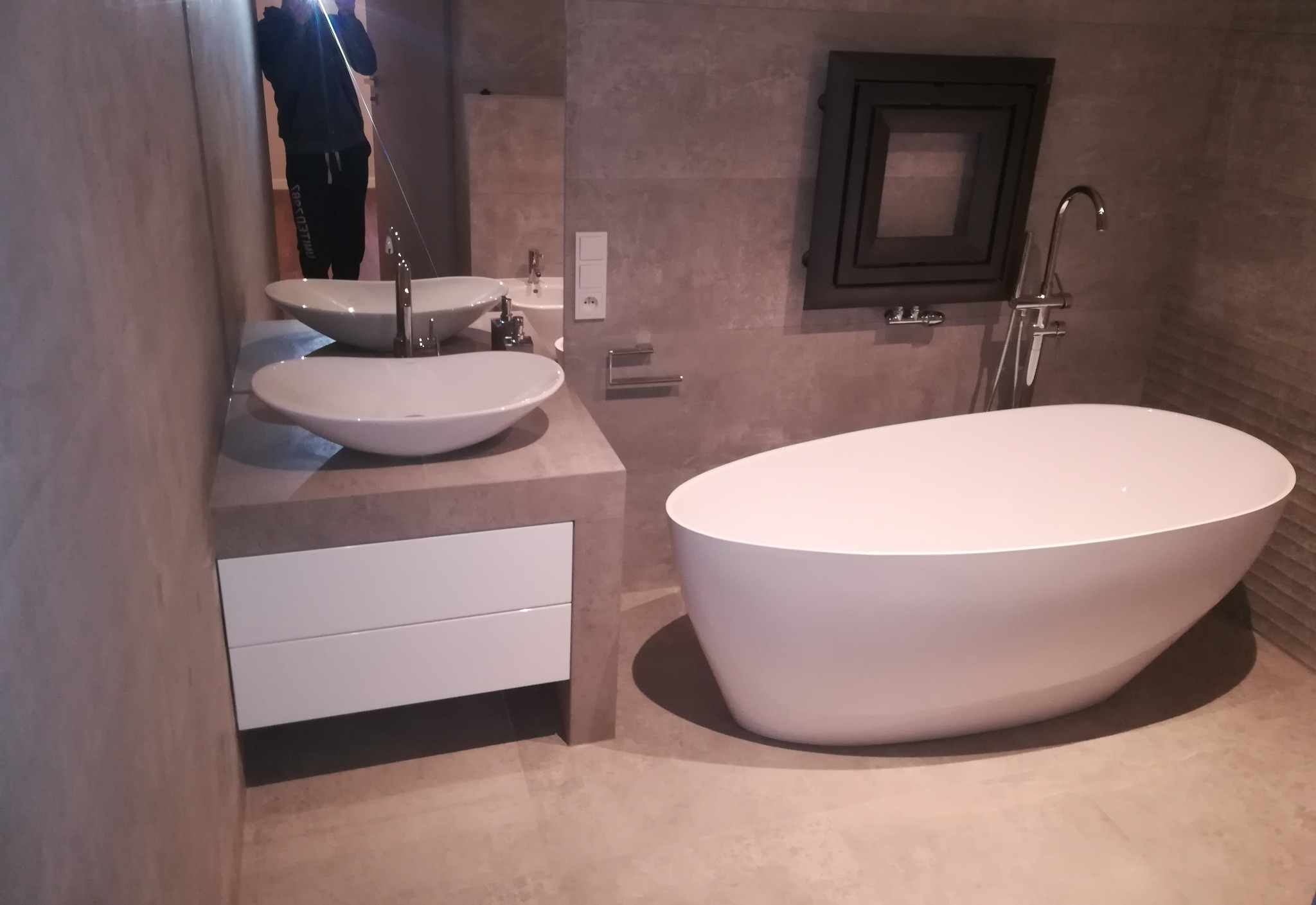 Nowoczesna łazienka: biała wanna wolnostojąca, umywalki nablatowe na szafce z szufladami, lustro z odbiciem osoby, szara płytka ścienna i podłogowa.