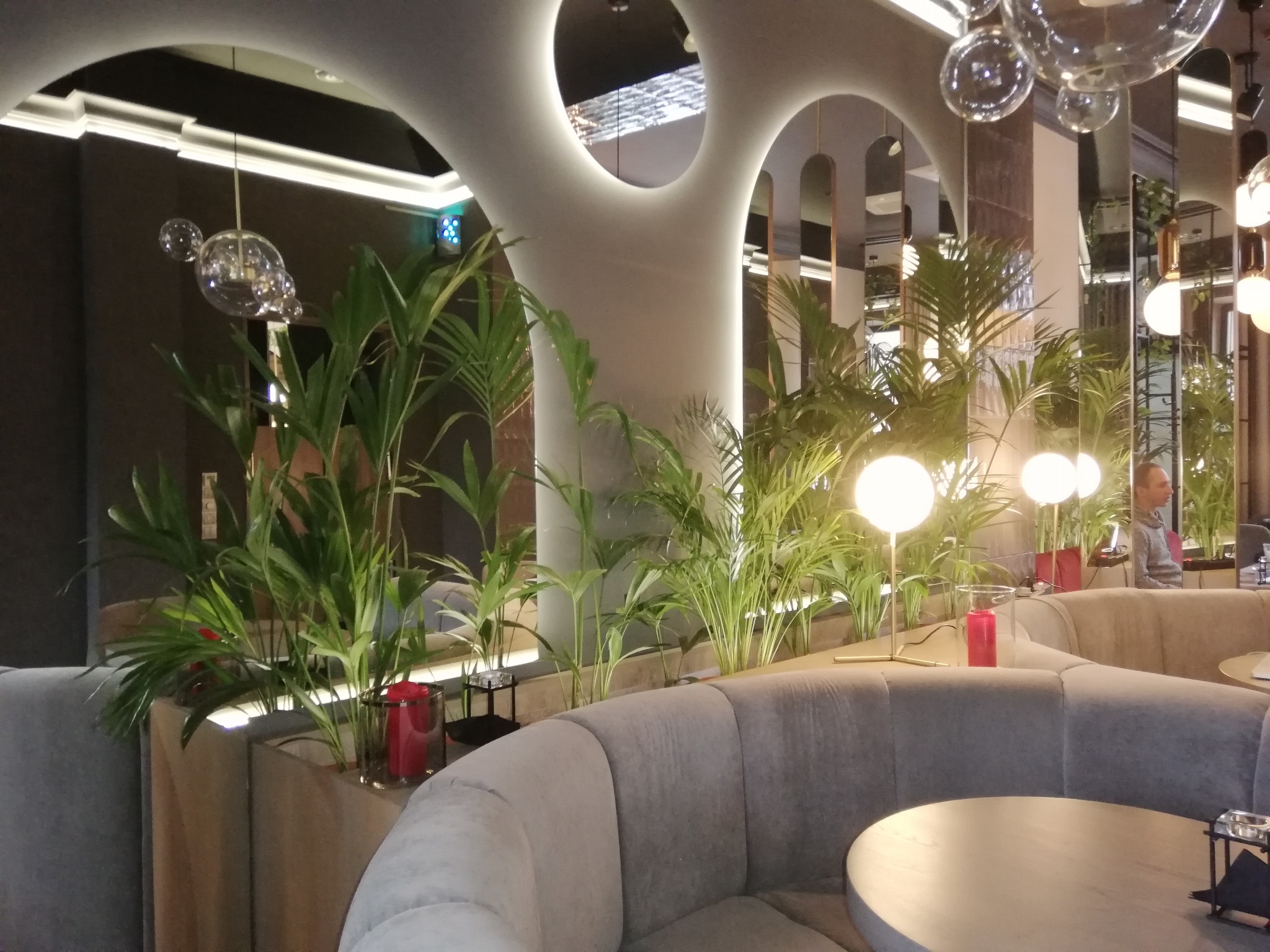 Nowoczesne wnętrze kawiarni z roślinami, okrągłymi stolikami i szarą tapicerką. Oświetlenie punktowe i dekoracyjne lampy kuliste. Minimalistyczny design.