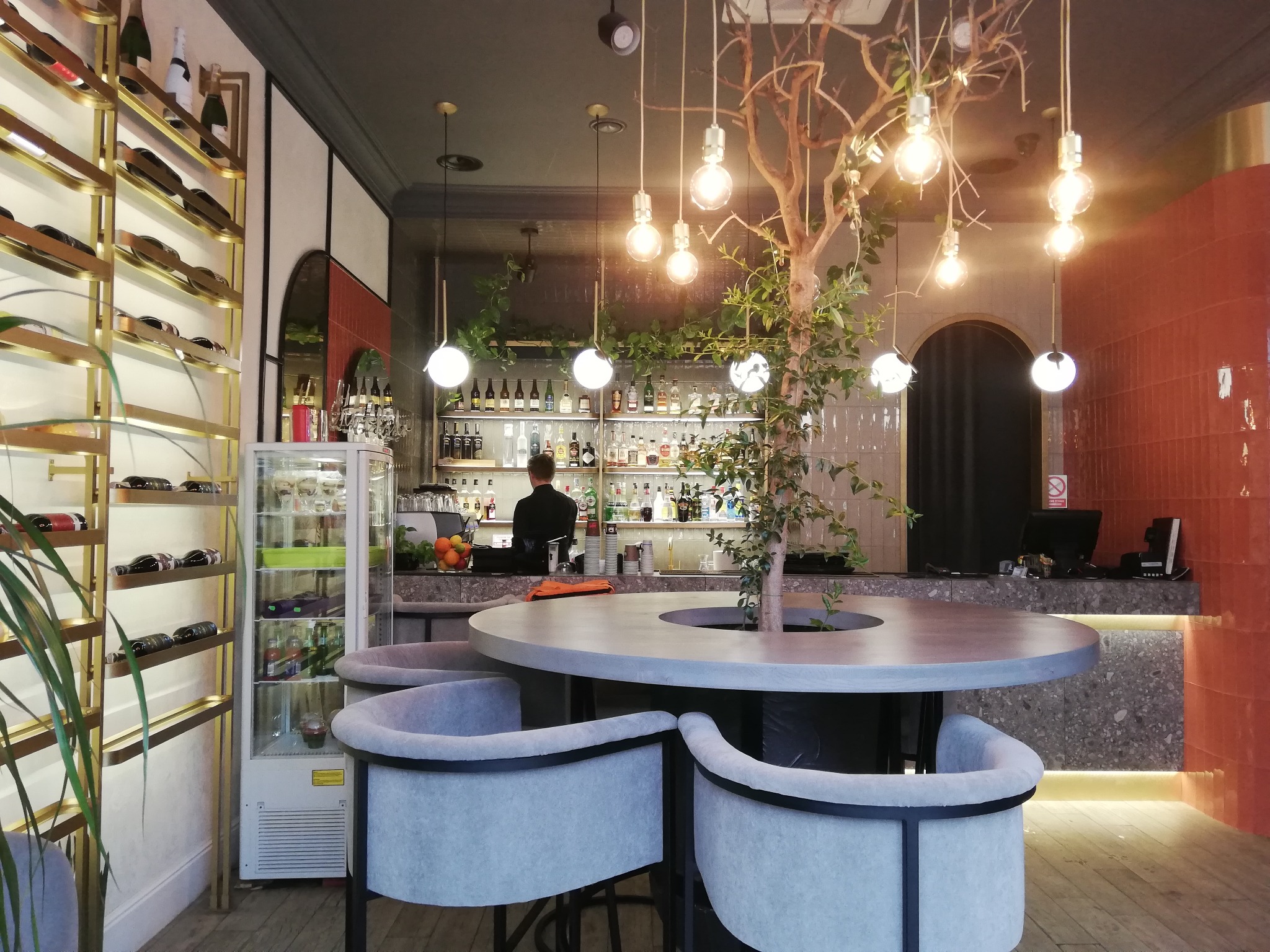 Stylowe wnętrze baru z okrągłym stolikiem otoczonym szarymi krzesłami. W tle bar z oświetleniem i dekoracyjnym drzewkiem. Na ścianie półki na wino.