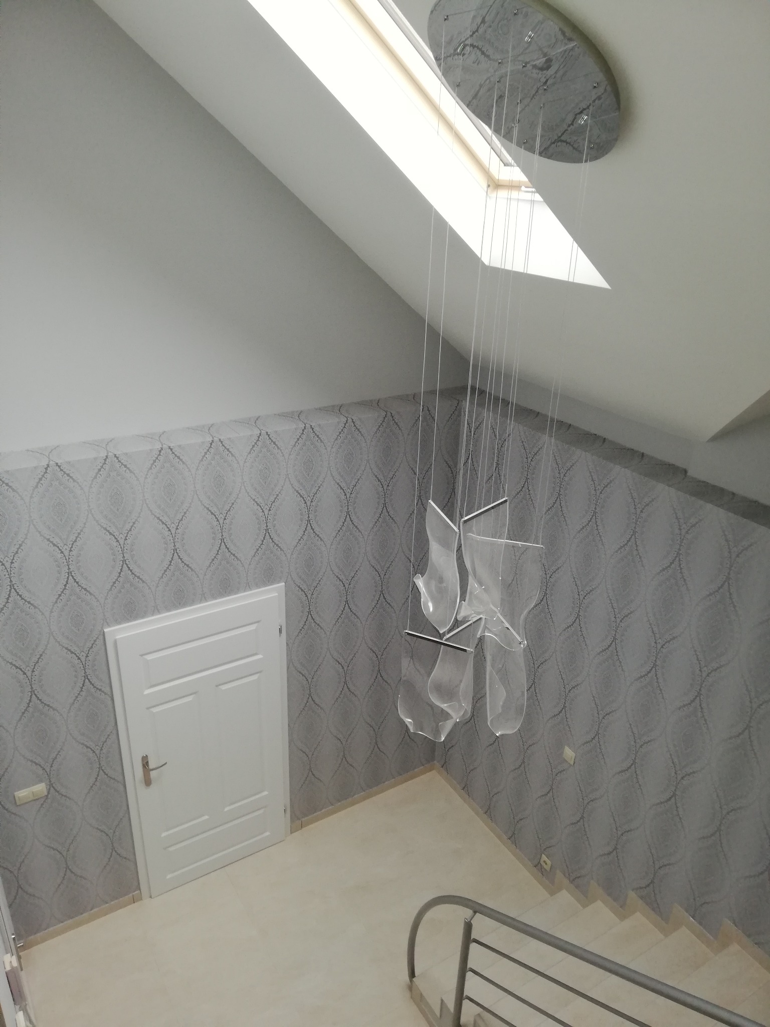 Klatka schodowa z szarymi tapetami w ornamenty, białe drzwi i schody z metalową barierką. Wisząca lampa z elementami z tworzywa sztucznego. Okno dachowe.