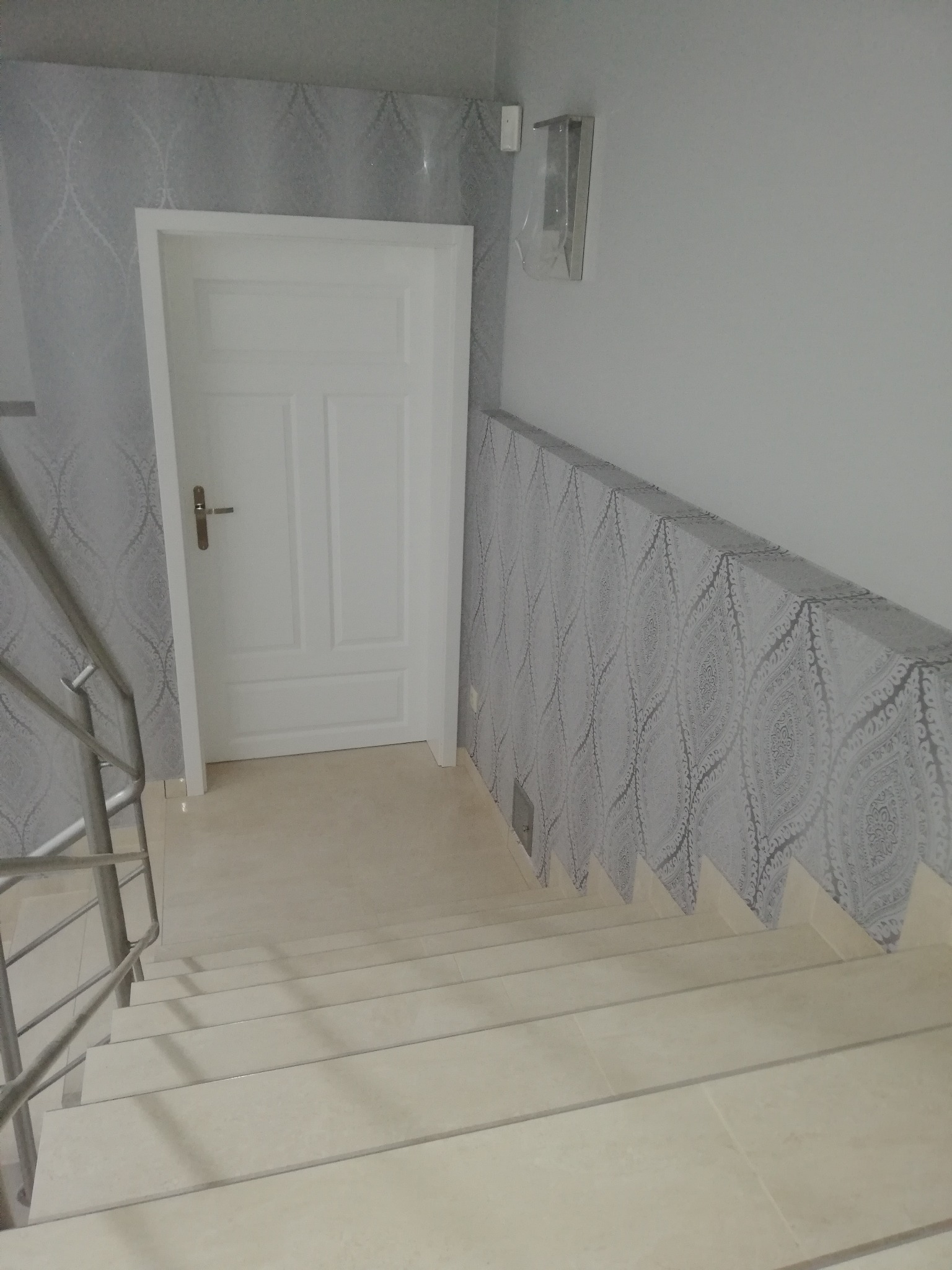 Wykończone schody z białymi stopniami i szarą balustradą. Ściany z szarą tapetą w ornamenty. Białe drzwi. Widok z góry na dół.