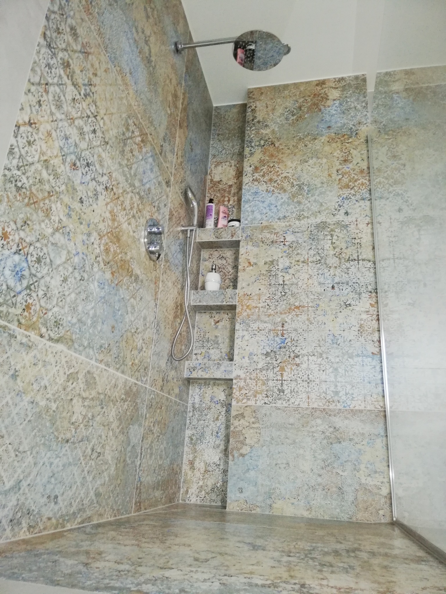 Wykończona łazienka z wielobarwnymi płytkami, kabiną prysznicową i półkami w ścianie. Bateria i deszczownica w kolorze chromu. Nowoczesne wnętrze.