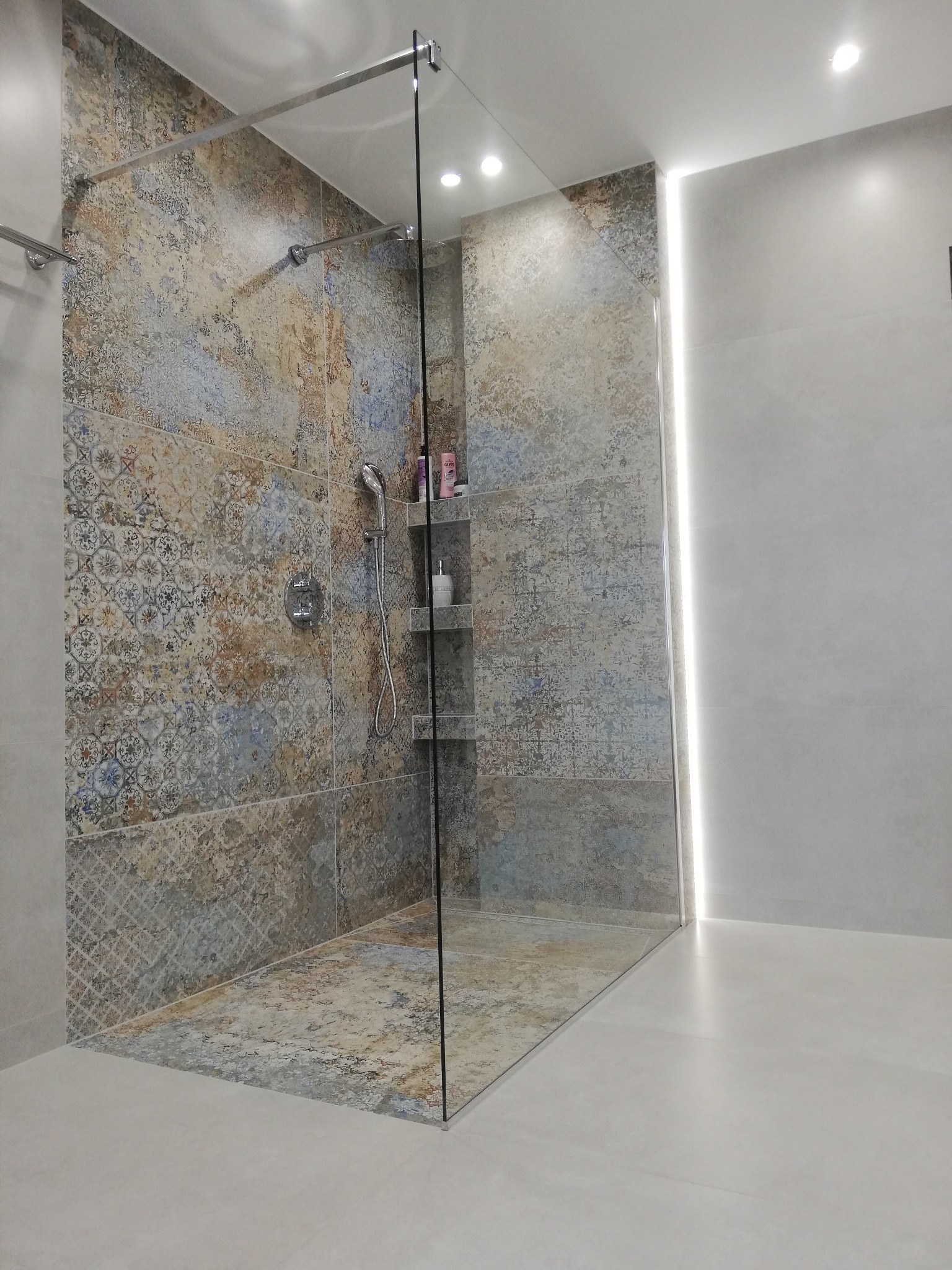 Nowoczesna kabina prysznicowa walk-in z dekoracyjnymi płytkami w odcieniach beżu i błękitu, półkami na kosmetyki i subtelnym oświetleniem LED.