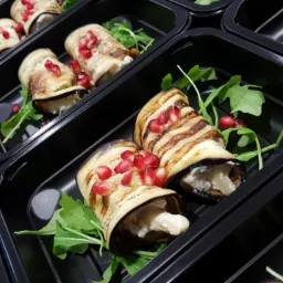 Grillowane roladki z bakłażana wypełnione serem, udekorowane granatem i rukolą, ułożone w czarnych pojemnikach cateringowych.