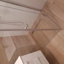 Zamknięta kabina prysznicowa z jasnymi, drewnopodobnymi płytkami w łazience w Rzeszowie. Widoczny biały słupek umywalki w rogu.