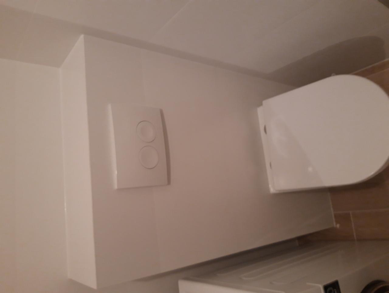 Biała zabudowa toalety podtynkowej z przyciskiem spłukującym na ścianie, widoczny fragment sedesu i pralki w łazience wykończonej płytkami imitującymi drewno.