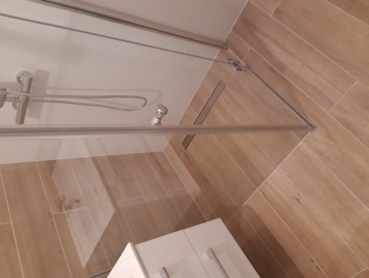 Zamknięta kabina prysznicowa z jasnymi, drewnopodobnymi płytkami w łazience w Rzeszowie. Widoczny biały słupek umywalki w rogu.