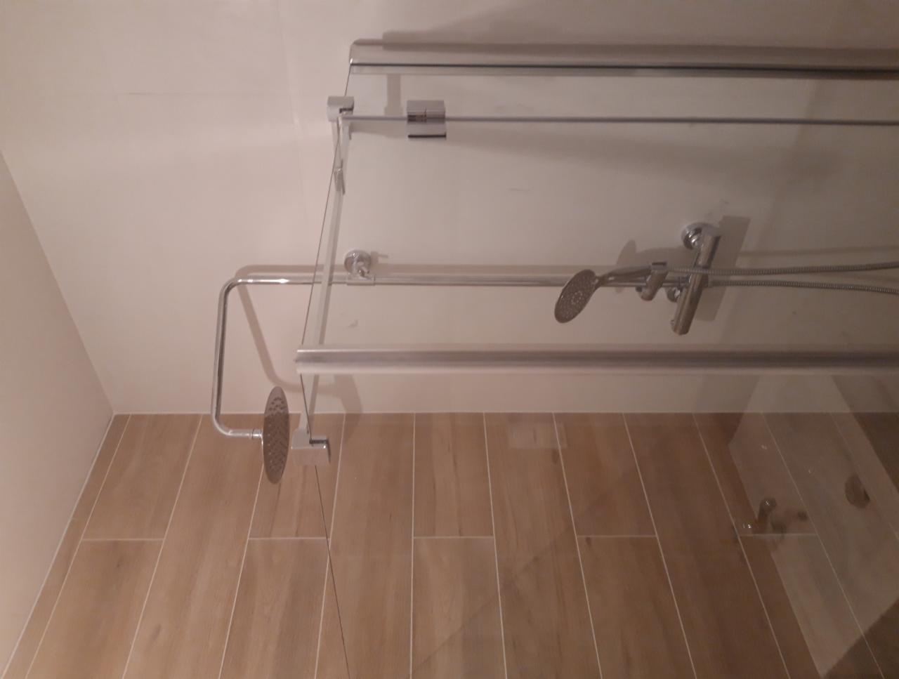 Szklana kabina prysznicowa z chromowanymi elementami, zamontowana na ścianie z beżowych płytek imitujących drewno, widok z dołu.