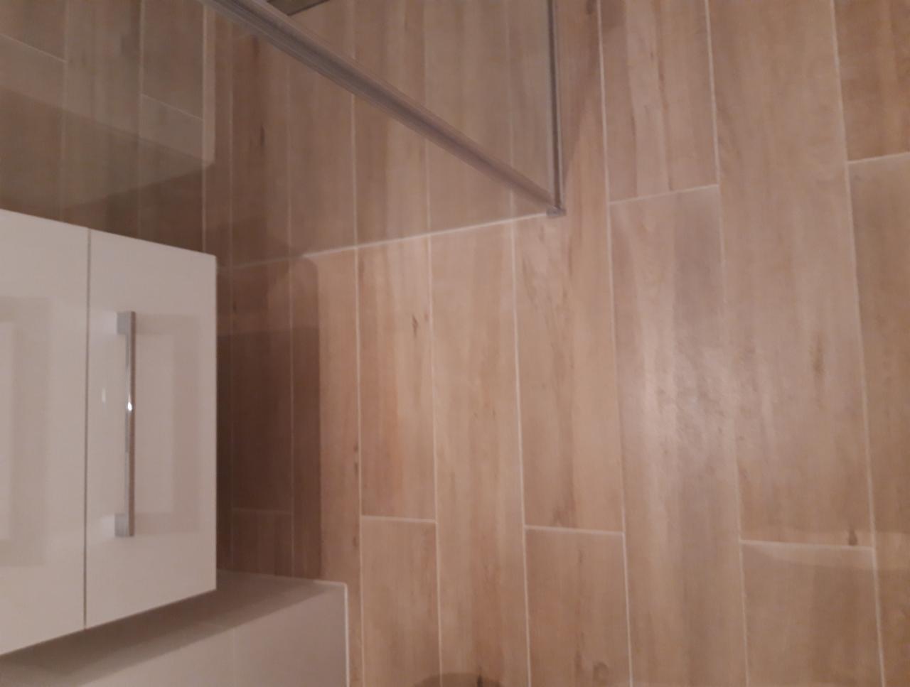 Jasna łazienka z kabiną prysznicową, beżowe płytki imitujące drewno na ścianach, biała szafka pod umywalką z metalowym uchwytem, widoczny fragment ramy kabiny.