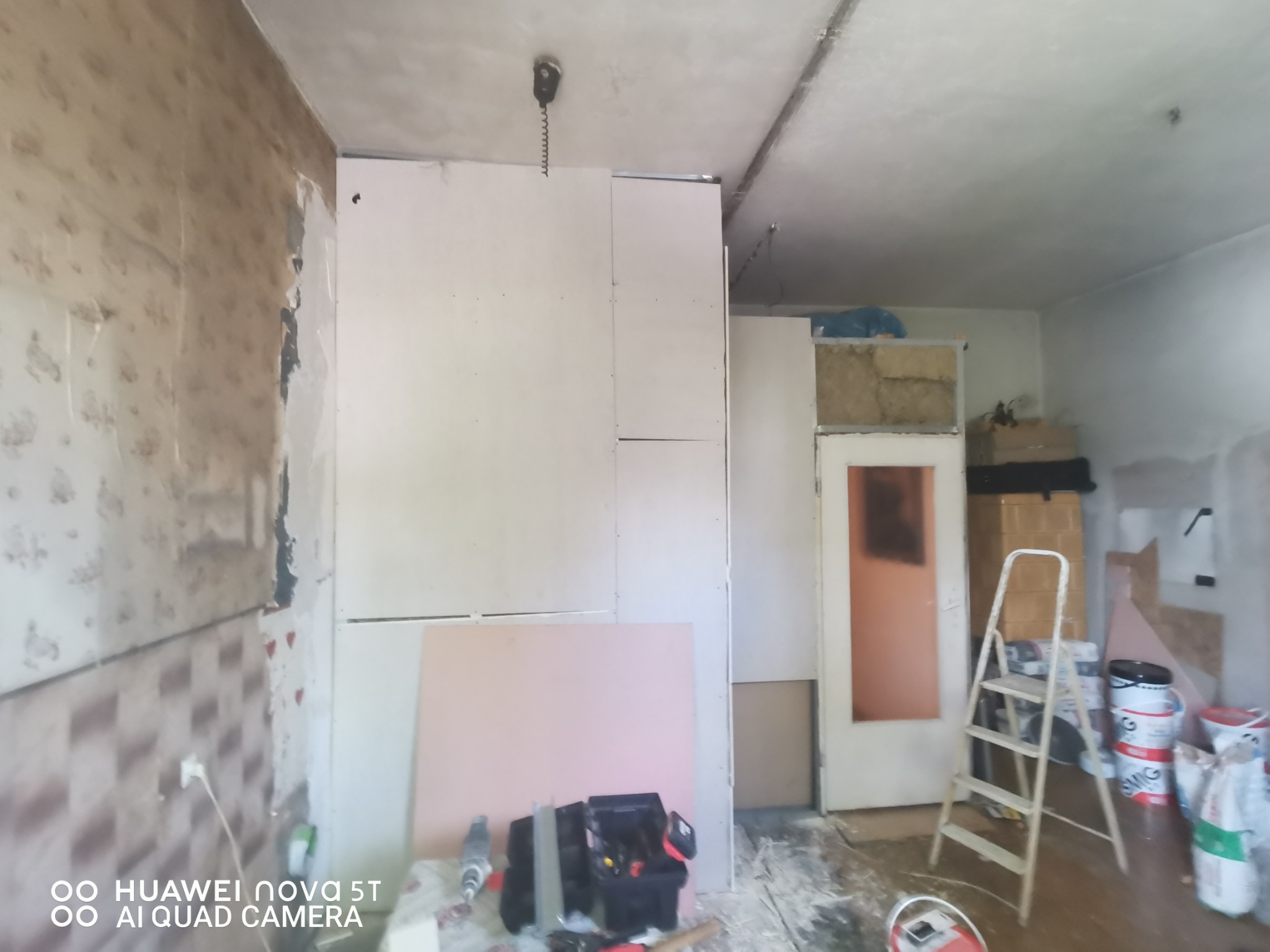 Pokój w trakcie remontu: widoczne płyty gipsowo-kartonowe na ścianie, stara tapeta częściowo zerwana, skrzynka z narzędziami, drabina, worki z materiałami budowlanymi, surowe wykończenie.