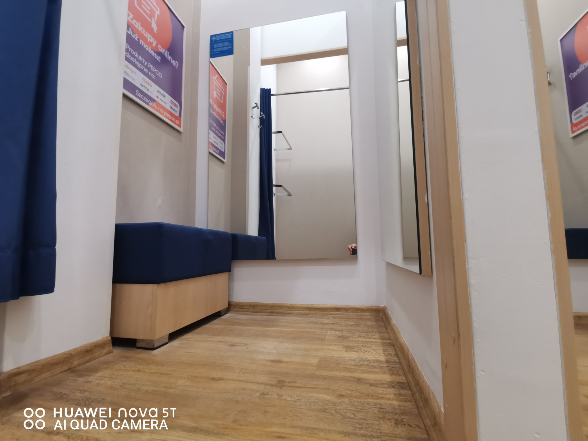 Wnętrze przymierzalni z drewnianą podłogą, lustrami i niebieską zasłoną, widok z dołu, na ścianie plakat reklamowy.