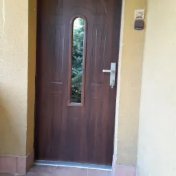 Brązowe drzwi wejściowe z podłużnym przeszkleniem, widok na zieleń ogrodu, srebrna klamka i zamek z kluczem, beżowa elewacja budynku.