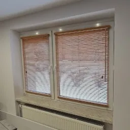 Dwa okna z zamontowanymi drewnianymi żaluzjami wewnętrznymi, widoczne podczas montażu, drabina oparta o ścianę, grzejnik pod oknem.