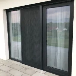 Przesuwne okno z ciemną ramą i moskitierą, widok z zewnątrz na taras z jasnej kostki brukowej, w tle domy i zieleń.