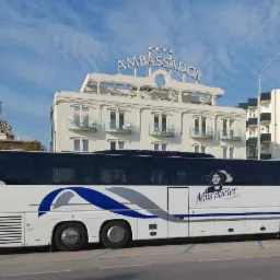 Autokar turystyczny z logo firmy 'Muszkieter' na tle hotelu Ambassador z czterema gwiazdkami, widok z boku, słoneczny dzień.