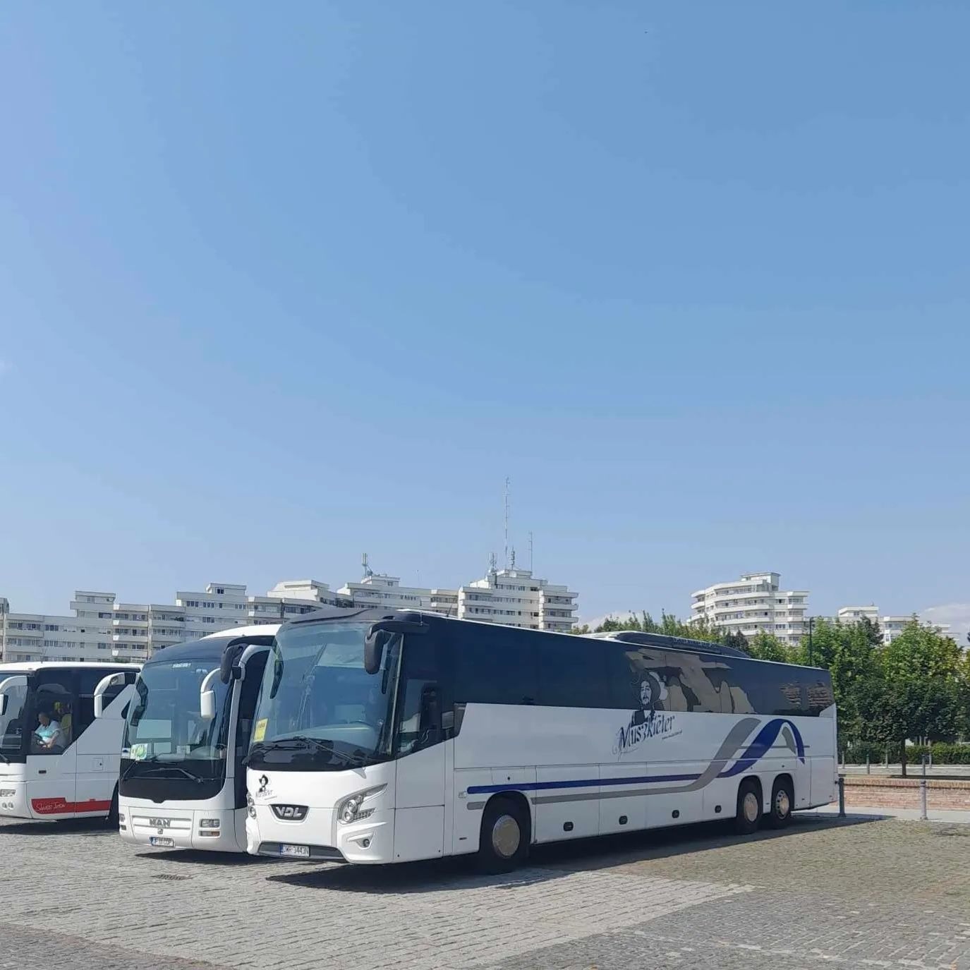 Flota autokarów turystycznych na parkingu z budynkami mieszkalnymi w tle, jeden z autokarów z wizerunkiem osoby i napisem 'Muszkieler'.