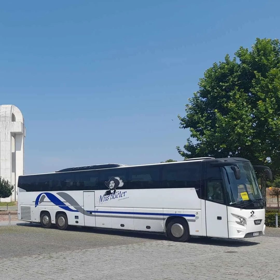 Biały autokar z grafiką muszkietera na boku, zaparkowany na brukowanym placu w słoneczny dzień.