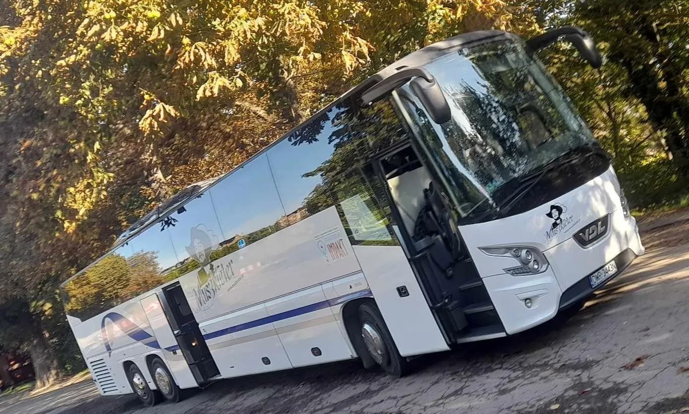Biały autokar turystyczny z otwartymi drzwiami, zaparkowany na tle drzew, z widocznym logo firmy 'Muszkater' na przedniej części.