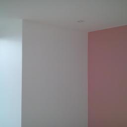 Usługi Remontowo-Budowlane Tomasz Majewski - Wnętrze pokoju z pomalowanymi ścianami w kolorach białym i pudrowym różu, widoczne okno z czarną ramą i fragment budynku na zewnątrz, minimalistyczne oświetlenie sufitowe.