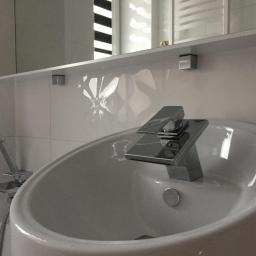 Usługi Remontowo-Budowlane Tomasz Majewski - Biała umywalka o zaokrąglonym kształcie z nowoczesną, chromowaną baterią i słuchawką prysznicową na tle białych, kwadratowych płytek z subtelnym wzorem. Nad umywalką widoczna biała półka z lustrem.