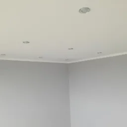 Wnętrze pokoju po remoncie: szare ściany, biały sufit z zamontowanymi punktowymi lampami i listwą dekoracyjną przy ścianie.