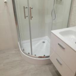 Usługi Remontowo-Budowlane Tomasz Majewski - Nowoczesna kabina prysznicowa narożna z chromowanymi detalami, umywalka z szafką w jasnym odcieniu, szare płytki podłogowe, minimalistyczny design łazienki.