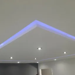 Nowoczesny sufit z podświetleniem LED w kolorze niebieskim, w kształcie geometrycznym, z zamontowanymi punktowymi lampami halogenowymi.