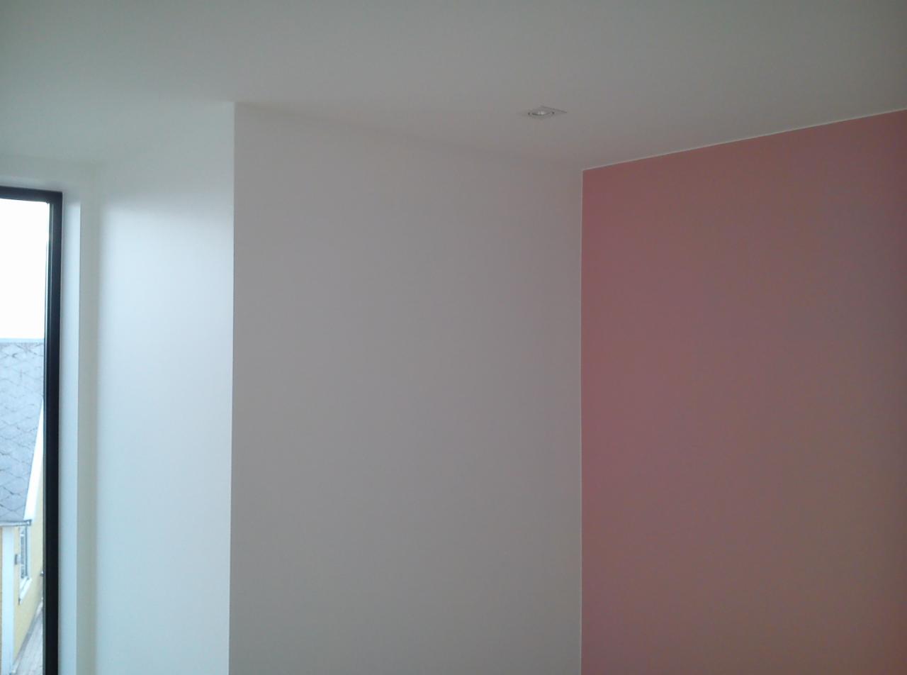 Wnętrze pokoju z pomalowanymi ścianami w kolorach białym i pudrowym różu, widoczne okno z czarną ramą i fragment budynku na zewnątrz, minimalistyczne oświetlenie sufitowe.