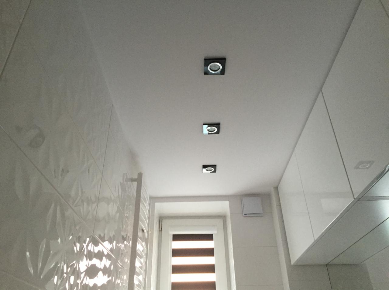 Nowoczesna łazienka z białymi, wzorzystymi płytkami na ścianie, trzema kwadratowymi lampami sufitowymi w czarnych obudowach, grzejnikiem drabinkowym i oknem z żaluzjami.