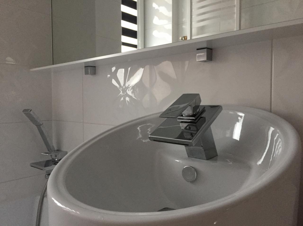 Biała umywalka o zaokrąglonym kształcie z nowoczesną, chromowaną baterią i słuchawką prysznicową na tle białych, kwadratowych płytek z subtelnym wzorem. Nad umywalką widoczna biała półka z lustrem.