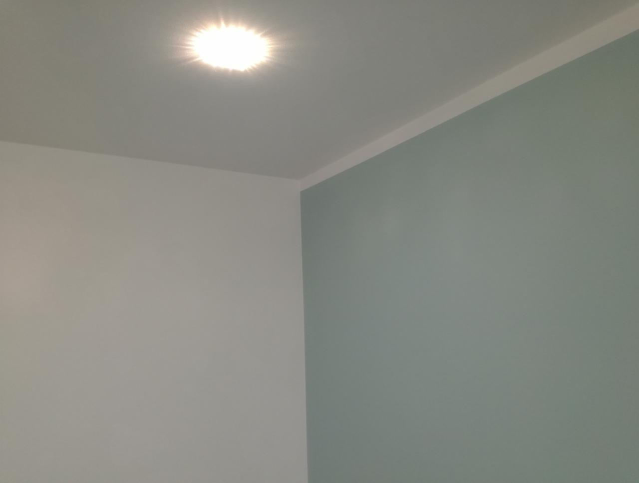 Świeżo pomalowany narożnik pokoju z jedną ścianą w odcieniu jasnej zieleni, drugą białą, oświetlony punktowym światłem w suficie.