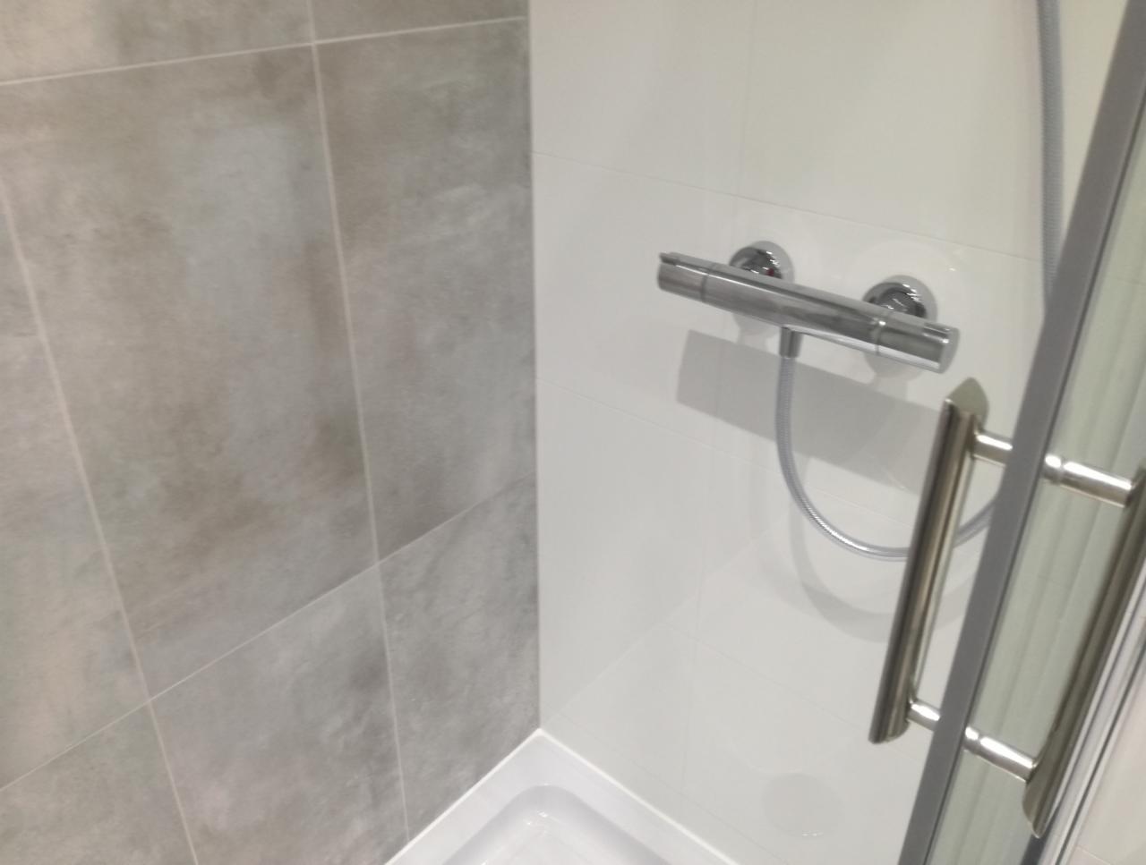 Fragment wykończonej kabiny prysznicowej z szarymi płytkami imitującymi beton na ścianie, białe płytki na pozostałych ścianach, chromowana bateria termostatyczna i uchwyt kabiny.