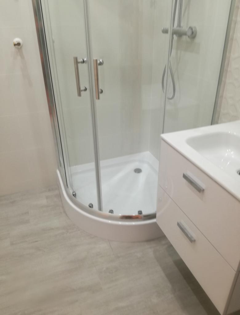 Nowoczesna kabina prysznicowa narożna z chromowanymi detalami, umywalka z szafką w jasnym odcieniu, szare płytki podłogowe, minimalistyczny design łazienki.