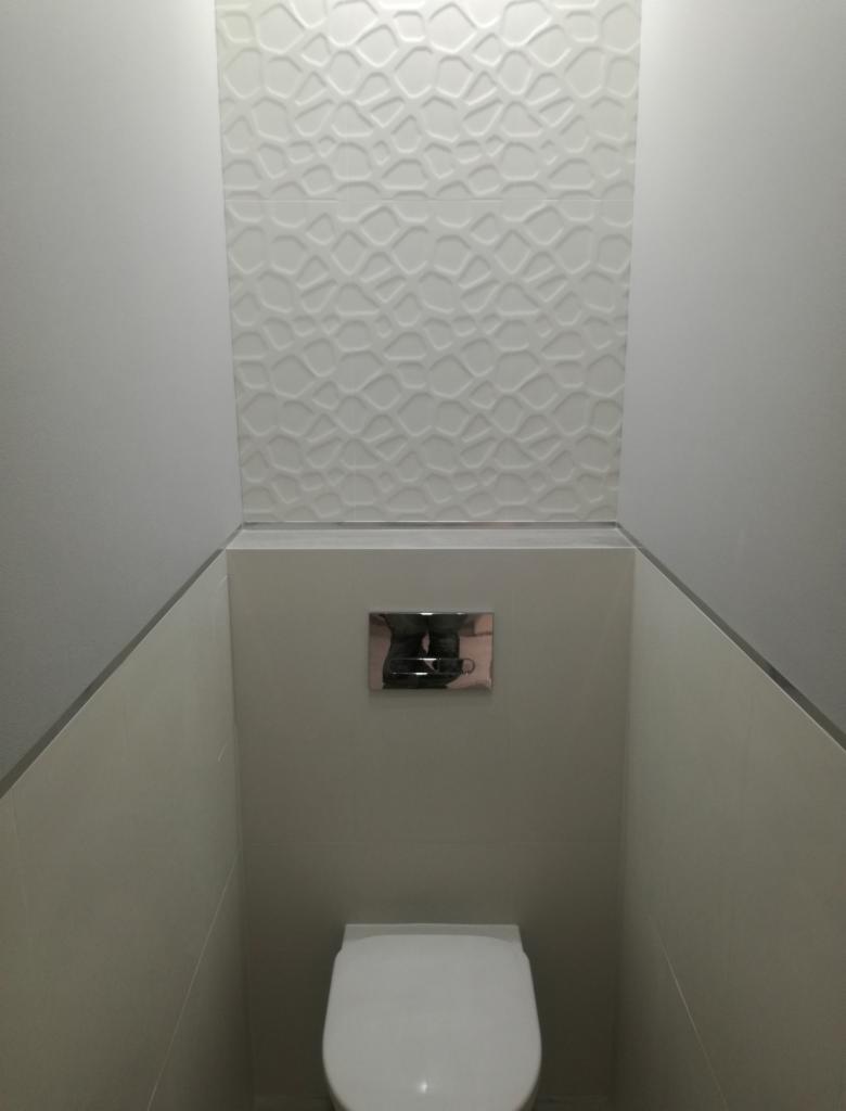 Wąska toaleta z białą miską wiszącą i geometryczną płytką dekoracyjną na ścianie, widoczny chromowany przycisk spłuczki.