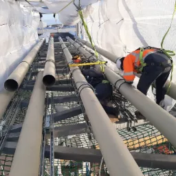 Pracownik w pomarańczowej kamizelce i kasku montuje rury na stalowej konstrukcji pod białym zadaszeniem, zabezpieczony uprzężą.