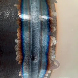 Detal spoiny na rurze, widoczne charakterystyczne przebarwienia termiczne i ślady po procesie łączenia metali. Powierzchnia rury częściowo pokryta ciemniejszym nalotem.