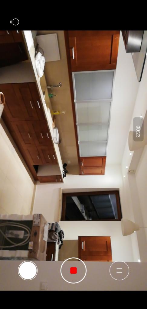 Kuchenne szafki w kolorze ciemnego drewna, zamontowane pod sufitem, z blatem i zlewem; widoczny fragment okna i lampy sufitowej.