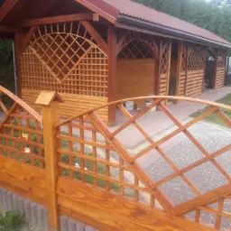 Drewniana altana z kratownicami i czerwonym dachem, widoczna przez ozdobne, drewniane ogrodzenie z różami w ogrodzie.