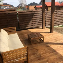 Zadaszony taras z drewnianą podłogą, drewnianymi słupami i poziomą, drewnianą balustradą. Widoczny drewniany stolik kawowy i sofa z białymi poduszkami. W tle zabudowa jednorodzinna.