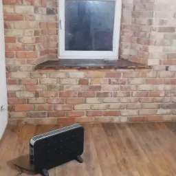 Pokój po remoncie z ceglaną ścianą z oknem i drewnianą podłogą, widoczny grzejnik elektryczny i przewody na podłodze.