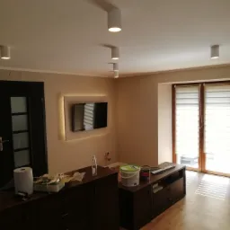 Beżowe ściany po świeżym malowaniu w salonie w Niemczy, z widocznymi puszkami farby i akcesoriami malarskimi na komodzie, oraz zamontowanym telewizorem z podświetleniem LED.